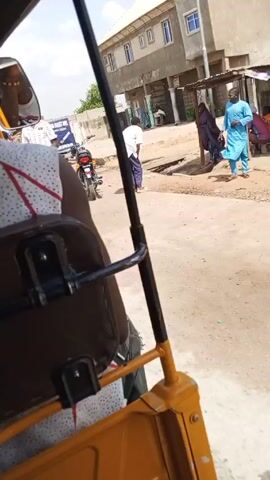 A yellow motorized rickshaw is moving along a paved road in what appears to be a town or village. The camera is positioned behind the driver, looking out the back of the vehicle.  The driver is wearing a white tank top with a red and black graphic design. The seat behind the driver is brown. Through the driver-side mirror, the driver's face is visible, wearing sunglasses and possibly a wristband. Another similar yellow rickshaw with "GATAN TATA" written on it is visible in front of the first rickshaw. A man in a white shirt and green pants stands on the side of the road. A motorcycle is parked nearby, and buildings are visible in the background. The sky is bright and sunny.