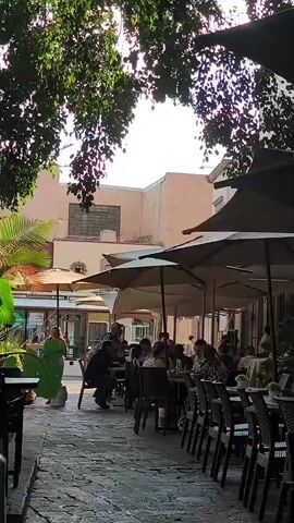 The video shows an outdoor cafe with several tables of people dining under umbrellas. The cafe is located on a stone-paved walkway lined with chairs. A young woman walks through the cafe carrying a tall glass. In the background, other people stroll by. The scene is partially shaded by trees.