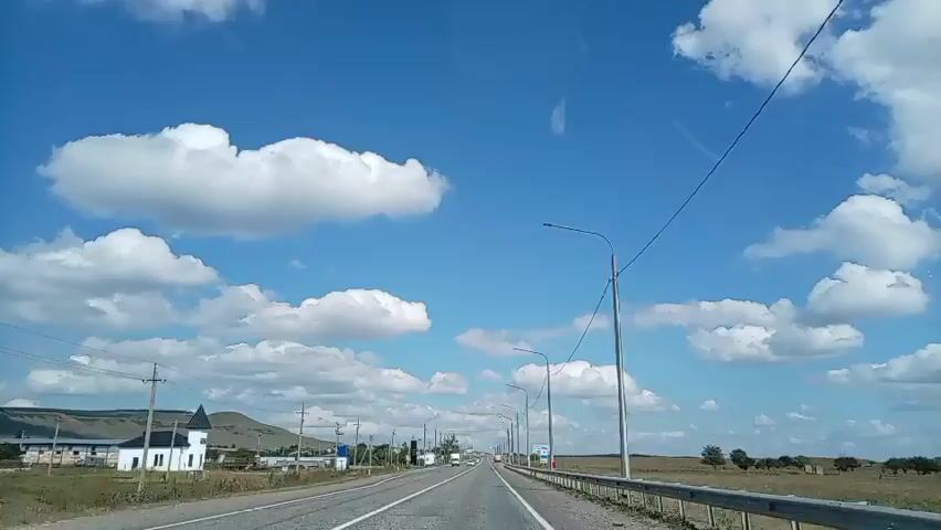 The video shows a view from a vehicle traveling on a highway. The road is paved with two lanes in each direction, separated by a solid white line. The sky is a vibrant blue with scattered white clouds. On the left side of the road, there are houses, some with pointed roofs, and trees. There are also power lines and utility poles running alongside the road. On the right side of the road, there is a metal guardrail and a vast, open landscape with dry grass and some scattered trees. Several cars and trucks are visible on the road, moving in the same direction as the camera. A signpost with Russian text is visible on the right side of the road, indicating a town or location. The overall impression is of a clear, sunny day with light traffic on a rural highway.