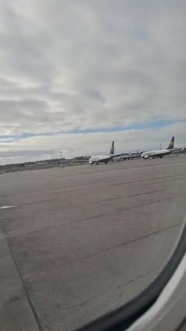 The video is taken from inside of a plane, looking out a window onto an airport tarmac. The sky is cloudy with patches of blue. Two Ryanair airplanes are parked on the tarmac, with the company logo visible on their tails. Other airport infrastructure, like towers, are visible in the distance. The camera is static and no people are visible in the video.