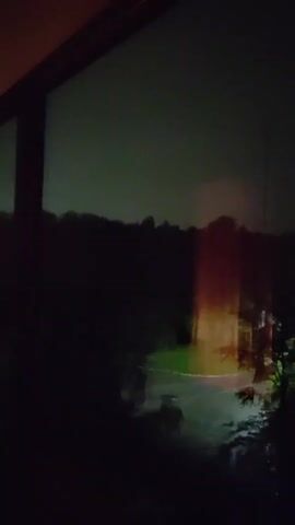 The video displays a view through a window at night or during a storm. The scene outside is dark and appears to be a natural landscape with trees and possibly a road. There are reflections on the window, some of which are blurry and indistinct. The lighting changes throughout the short clip, with periods of darkness and brief flashes of light, suggesting either lightning or artificial lights. The presence of water droplets on the window indicates that it is raining.
