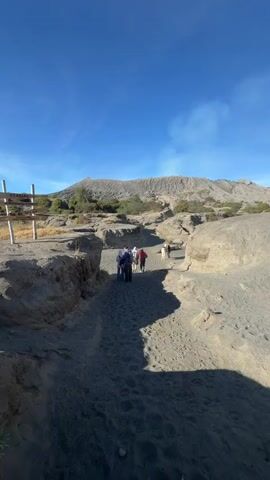 A group of people walk along a sandy path toward a volcano. The path is narrow and enclosed by rocky terrain. The sky is clear and blue. A signpost is visible to the left of the path. The volcano appears to be active, with a plume of smoke rising from its summit.