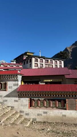 The camera pans right revealing a Tibetan monastery with a red roof and a wall adorned with prayer wheels. The backdrop features rugged mountains, and snow-capped peaks are visible in the distance. The sky is clear and blue. In the foreground, a sandy clearing has two people visible, one kneeling and possibly taking a picture, and another person standing and looking at the scenery. A third person is walking with a backpack on. The scene conveys a sense of tranquility and majestic natural beauty.