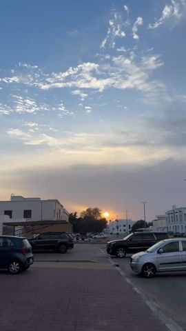 The video shows a parking lot at dusk, with the sun setting behind a tree. The sky is partly cloudy, and the light is soft and golden. Several cars are parked in the lot. The buildings in the background are white. The video ends abruptly and is accompanied by a song in another language.