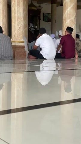 This media file captures a scene inside a mosque in Rhieng Blang, Indonesia. The foreground features a highly reflective tiled floor, mirroring the pillars and people above. Several individuals, all men, are seated on the floor, facing towards the front of the mosque where a pulpit is visible. They appear to be engaged in prayer or a religious gathering.

The subjects are dressed in traditional Islamic attire. One man in the center wears a white shirt and black pants, his back to the camera. To his right, another man is in a maroon shirt and black pants. Other individuals are partially visible, some wearing white skullcaps. Behind them, a preacher or religious leader stands at the pulpit.

The setting is clearly a mosque, indicated by the presence of the pulpit and the formal attire of the attendees. The pillars are decorative, with a marble-like finish. The time of day is suggested by the lighting, which appears to be natural, possibly late morning or afternoon, but not definitively. There are no explicit weather cues.

A visible piece of text appears on a digital display in the background, showing "12.58". The overall emotion conveyed is one of reverence and communal worship. The reflective floor adds an artistic and almost surreal quality to the image, emphasizing the solemnity of the occasion.