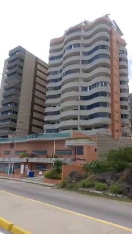 The video shows a view of two tall buildings from a street. The building on the left is a rectangular, grey structure with many windows and balconies. The building on the right is taller and has a wavy, curved facade with numerous balconies. In front of these buildings is a lower, brick structure with several windows and a garage door. A green and white sign with text is visible on a pole in front of the wavy building. There is also a blue tent set up on the sidewalk. The street in front of the buildings has yellow and white markings. The sky is overcast. The video appears to be shot from a moving vehicle.
