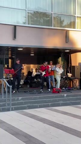 A band performs on an outdoor stage in front of a building. The band consists of a bassist on the left, a guitarist in the center, a keyboardist behind them, a guitarist/vocalist next to the keyboardist, and a female vocalist on the far right. The bassist is wearing a black t-shirt and black pants. The guitarist in the center is wearing a red t-shirt and black pants, and is playing a red electric guitar. The keyboardist is wearing a white shirt and brown pants, and is playing a keyboard. The other guitarist/vocalist is wearing an orange shirt and brown pants, and is holding a music stand. The female vocalist is wearing a light-colored outfit and is holding a microphone. There are several musical instruments and equipment on the stage, including drums, amplifiers, and speakers. The stage is set up on steps leading up to the building's entrance. The building has large windows and an awning. The lighting suggests it is late afternoon or early evening.
