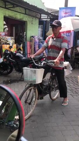 A man wearing a red and white striped polo shirt, black pants, a baseball cap, and flip-flops stands next to a bicycle with a white basket on the handlebars. He is holding onto the handlebars, appearing to be either preparing to ride or having just dismounted. The bicycle is dark colored with visible spokes on the wheels. Behind the man, a black motorcycle is parked. The environment appears to be an outdoor market or street, with stalls selling various items, including what looks like rolled-up fabrics or rugs with colorful patterns. A green building is visible on the left, and part of a blue and white sign is visible on the right. The ground is paved with interlocking bricks. The video is shot from a low angle, possibly from another vehicle or a person at a similar height.