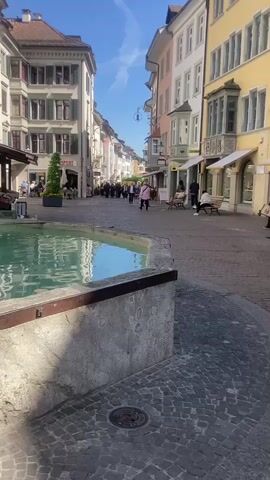 The video showcases a vibrant pedestrian street in Schaffhausen, Switzerland, on a sunny day. In the foreground, a large, octagonal stone fountain with clear, greenish-blue water dominates the lower left of the frame. The fountain features a central statue and a beautifully arranged circular flower bed, bursting with purple, yellow, and white pansies, positioned just above the water's surface.

The street itself is paved with cobblestones, and lined on both sides by multi-story buildings with traditional European architecture, featuring numerous windows and colorful facades in shades of white, yellow, and beige. People are seen strolling leisurely along the street, some gathered near storefronts or sitting on benches. A wooden market stall is visible near the fountain.

The weather is bright and clear, with a vivid blue sky overhead, indicating daytime. Visible text includes "WALDER" on a shop sign and "SPORT" on another. The overall atmosphere appears peaceful and lively, capturing a typical day in this charming Swiss town.
