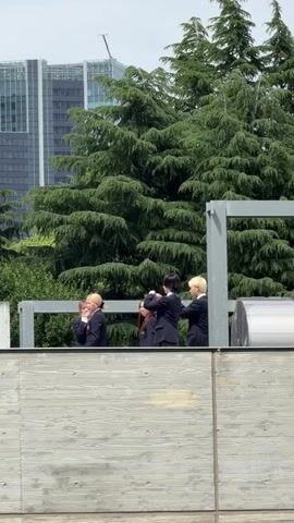 Five individuals in dark suits stand on a rooftop or elevated platform. Two individuals on the far left are interacting, one with blonde hair and the other with dark hair. To their right, another individual with dark hair stands. Further to the right, two individuals are facing each other, one with dark hair and the other with blonde hair. Behind them are lush green trees and a tall building. The scene is captured during daylight.