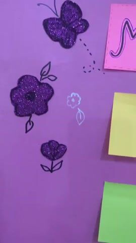 A framed poster board with a purple background is decorated with various elements. At the top, large, glittery purple letters spell out "CONGRATS!". Below this, a pink banner features the words "Muzzamil Ahmed" written in cursive. To the right of the name, a small pink square has the date "17 SEP 2026" written on it. Scattered across the poster are several glittery purple butterflies and flowers, some with black outlines. There are also some smaller, hand-drawn white flowers and leaves. Several sticky notes are attached to the board: one on the left with handwritten text in a foreign language and the English words "Congratulations"; one to its right is blank and yellow; another sticky note below the first is orange with handwritten text; and a final yellow sticky note is to the right of the orange one and is blank. The overall impression is a celebratory handmade poster.
