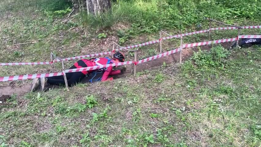 The video shows two people crawling through a narrow dirt trench in a wooded area. The trench is marked with red and white striped tape strung between poles. The person in the foreground is wearing a red jacket, black pants, and a helmet. They are crawling on their hands and knees. The second person, further down the trench, is wearing a black outfit and a red helmet, also crawling. The ground around the trench is covered in grass and some dirt. Trees and foliage are visible in the background.