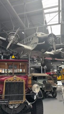 The video shows a museum exhibit of vintage vehicles and an airplane. In the foreground, a dark red truck is visible from the front. Behind it, a lighter colored truck, possibly green or gray, is seen. Further back and suspended from the ceiling is a black and silver airplane with two propellers on each wing. The airplane has "Lufthansa" and "TEMPELHOF" written on its side. The environment appears to be a large indoor space with a high ceiling and natural light coming from skylights. There are other exhibits and displays visible in the background, as well as a few people in the distance. The camera is static and the footage is of good quality.