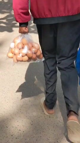A person in a red jacket and black pants is walking on a sidewalk. They are carrying a bag of eggs in their left hand and a large blue plastic bag in their right hand. The person is wearing slippers. The sidewalk is gray and there are shadows cast on it. In the background, there is a brown sofa and a few people standing near it. There are also cars and motorcycles passing by on the street.