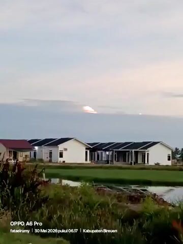 The video shows a line of single-story houses against a muted sky, with a small body of water and grassy vegetation in the foreground. The houses are predominantly white with dark gray roofs and visible air conditioning units. The sky is light, and the sun, obscured by clouds, creates a bright spot on the horizon. Information at the bottom of the frame indicates the video was taken with an OPPO A6 Pro on May 30, 2026, at 6:17 in Kabupaten Bireuen. Sounds of crickets or other insects can be heard in the background.