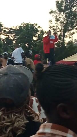 A man in a red jacket stands on a stage and addresses a crowd of people, who are seated on the ground. The man is holding a book and speaking into a microphone. The crowd appears to be composed mostly of women and children. The setting is outdoors, with trees and a sunset visible in the background. The video is shot from a low angle, looking up at the stage. The image is slightly blurry and the colors are muted, suggesting it was taken in low light conditions.