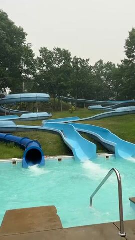 A young person in a pink swimsuit slides down a blue water slide into a pool of turquoise water. Behind them, more blue water slides wind through a grassy, tree-lined area under an overcast sky. Another person in a dark swimsuit is visible in the pool, to the left. The person on the slide splashes as they enter the water. The video continues as the first person surfaces in the pool, followed shortly by the second person.