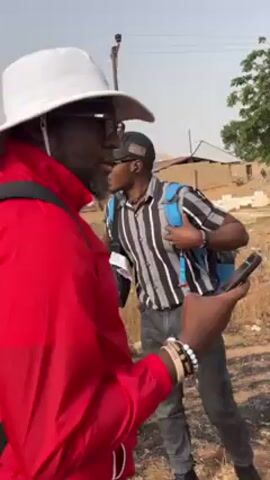 Here's a description of the video:

The video shows three people standing outdoors. The person closest to the camera is wearing a red jacket, a white hat, and glasses. They have a backpack on and are holding a phone. Another person, standing next to them, wears a black and white striped shirt, a black cap, glasses, and a blue backpack.  A third person, with braided hair, wears a black jacket. They are holding and looking at their phone. The background includes sparse vegetation and some buildings. The scene appears to be taking place during daylight hours. The three individuals appear to be interacting.