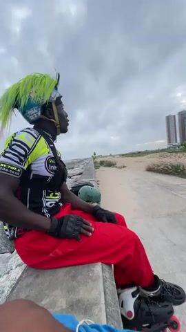 A man wearing roller skates, a helmet, and a neon green mohawk is sitting on a concrete ledge outdoors. He is wearing a black and yellow shirt with red pants and black gloves. Another person is visible in the foreground, also wearing roller skates. In the background, there is a beach, a paved path, and several tall buildings under a cloudy sky. The man on the ledge appears to be talking or singing. He then stands up and begins to skate away, followed by the other person. The man in the black shirt and purple pants skates towards the camera and then turns away.