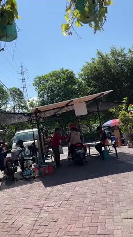The video shows a small outdoor market or roadside stall on a sunny day. In the foreground, there is a brick-paved area. Behind this, a covered structure with a white awning serves as a shelter. Underneath the shelter, several people are present. One person in a red shirt stands to the left, appearing to interact with someone on a scooter. Another person, wearing a light brown jacket, is on the scooter, facing away from the camera. To the right, another individual is seated on a stool. Various objects are visible, including motorcycles parked to the left of the shelter, bags, and what appear to be merchandise or supplies. In the background, a white van is parked, and further beyond, trees and a power transmission tower are visible under a clear blue sky. Green foliage hangs from above, partially obscuring the view of the sky. The overall atmosphere suggests a casual, possibly commercial, outdoor setting.