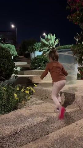 A young girl with blonde hair tied in a ponytail runs up a set of outdoor stairs at night. She is wearing a brown long-sleeved shirt and white patterned leggings with pink shoes. The stairs are made of a speckled material and are surrounded by plants and large decorative planters. The sky is dark, and streetlights illuminate the scene.