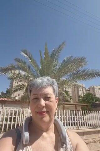 A selfie-style medium shot video features a middle-aged woman with short, gray hair, wearing a white sleeveless top and a gray neck fan, talking to the camera outdoors on a bright, sunny day. She is smiling and speaking in a language other than English. In the background, there is a large palm tree, white fence, and buildings under a clear blue sky. The video has no cuts or special effects and is approximately 5 seconds long.