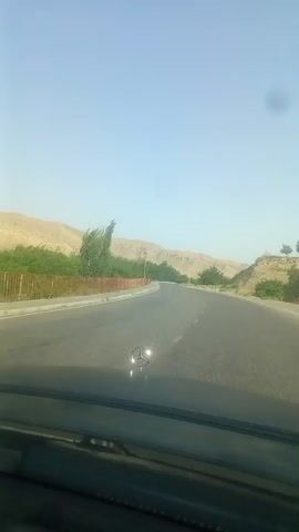 The video shows a view from inside a car driving down a paved road. The road curves to the right, flanked by a low reddish-brown fence on the left, behind which are trees and a sloping, arid hillside. Further in the distance, more hills are visible under a clear, pale blue sky. The hood of the car, with a Mercedes-Benz emblem, is visible in the foreground. Power lines stretch across the sky. The time stamp indicates the video is just a few seconds long.