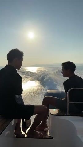 Two individuals, silhouetted against a bright sun and clear sky, sit on the stern of a moving boat. The boat is creating a significant wake of white water. The person on the left is wearing a black shirt and shorts, with their legs extended. The person on the right is wearing a dark shirt and shorts and is looking forward, with one hand near their face. The environment appears to be a body of water, possibly the ocean, with distant land visible on the horizon. The sun is high in the sky, indicating daytime.