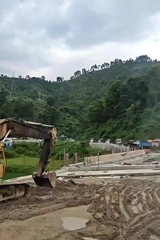 The video shows a construction site in a mountainous area. In the foreground, there is mud and construction materials, including rebar and wooden planks. An excavator is partially visible on the left. In the middle ground, there is a partially constructed building with concrete foundations and vertical rebar. A road with several vehicles, including cars and buses, runs along the base of a green, tree-covered mountain in the background. The mountain has terraced fields and scattered houses. The sky is overcast. The camera pans slightly to the right.