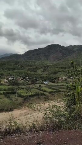 The video captures a panoramic view of a rural village nestled amidst lush green hills under a cloudy sky. The village appears to consist of scattered houses, some with red roofs, spread across the landscape. Terraced fields with green vegetation are visible on the slopes. A dry riverbed or a stream with sandy banks runs through the foreground. The overall scene suggests a remote, natural setting.