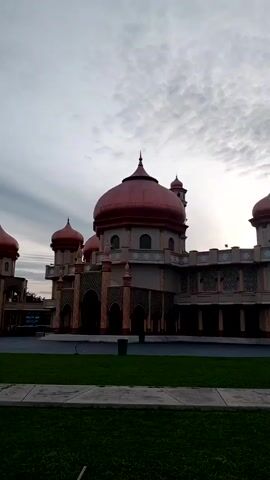 The video depicts a large building with multiple red domes, likely a mosque, set against a cloudy sky. The building is beige with intricate patterns and arched openings. A green lawn is in the foreground, separated from the building by a paved area. There are people sitting on the lawn and some bushes near the building. The sun appears to be setting behind the building, creating a silhouette effect.