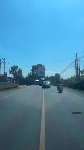 The video shows a road in a rural area on a sunny day. A car and a scooter are driving on the road. There are trees and bushes on the sides of the road, and power lines are visible in the background. The road is paved and has a yellow line dividing the lanes. The sky is clear and blue.
