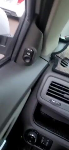 The video shows a view from the passenger seat of a car on a highway. The car is moving forward, and the mountains are visible on the right side of the road. Several other vehicles are on the highway, including a white car in front, a dark-colored car, and a truck in the distance. The mirror on the side of the car shows a person's head and shoulders, suggesting the video is being filmed by the passenger. The sky is clear, and the sun appears to be setting, casting a warm light on the scene.
