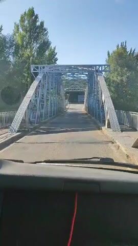 The video begins from the perspective of someone inside a vehicle, looking forward. The vehicle is on a road that leads onto a metal truss bridge. The bridge has a gray, weathered appearance, with visible structural beams and cross-bracing. The road surface on the bridge appears to be asphalt and slightly wet. Trees and vegetation are visible on either side of the bridge and in the background, suggesting a rural or semi-rural environment. The sky is clear and blue, indicating daytime. The vehicle appears to be moving forward onto the bridge. The video is short and shows the approach to and the beginning of crossing the bridge.