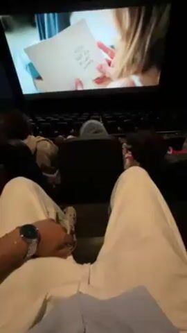 Here is a description of the video:

The video shows a first-person perspective of a person in a movie theater watching a film. The person is sitting with their legs stretched out, wearing white pants, a blue shirt, and a watch. In the seats in front of them are two other people. The theater is dark, with rows of empty seats. On the movie screen, various clips are showing a young woman reading. At one point, the screen displays the dates "July 04, 2025" and then "September 04, 2024."