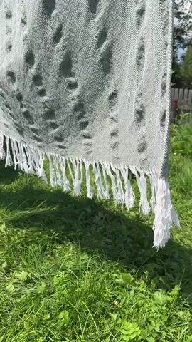A close-up shot of a light green and white woven blanket with a fringe is shown outdoors. The blanket is hanging and appears to be drying in the sun. Green grass and some foliage are visible in the background. The sun is casting shadows on the blanket, creating a textured effect. The blanket has a herringbone pattern.