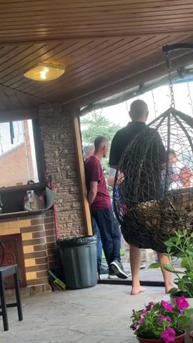 The video shows a group of people on a covered patio. In the foreground, a large hanging rattan chair is visible, partially obscuring the view of the people behind it. Several potted flowers, including pink and purple petunias, are arranged on the patio floor.

To the left, there is an outdoor kitchen area with a brick counter and a sink. A dark gray trash can and cleaning supplies are near the counter. A window looks out onto a brick building.

The people are standing and interacting. One man in a black t-shirt is facing away from the camera, and another man in a maroon t-shirt is leaning against a stone wall. Two other people are partially visible in the background, one in a blue shirt. The floor of the patio appears to be made of stone tiles. The ceiling is made of wooden planks, and there is a light fixture attached to it. The overall atmosphere suggests a casual outdoor gathering.