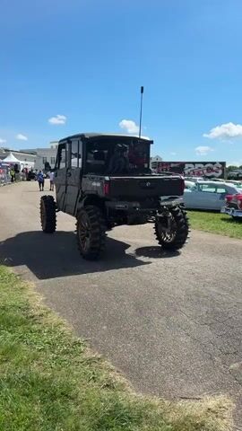 A black, modified off-road vehicle with large tires is shown from the rear driving slowly down an asphalt road. The vehicle has a crew cab and an open bed. In the bed, there are several large, black, tubular objects attached to what appears to be a rack. A black antenna extends vertically from the roof of the cab. The vehicle is outdoors on a sunny day. To the left of the road, there is grass and some tents and people are visible in the distance. To the right of the road, there are several classic cars parked on the grass. The background includes a white building with a mansard roof.