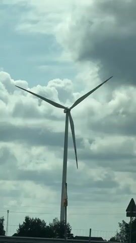 The video shows a wind turbine spinning against a cloudy sky. The turbine is white, with three blades tipped in red. It stands tall in front of a line of trees, with power lines also visible in the background. The camera appears to be inside a moving vehicle, as road barriers can be seen in the foreground.