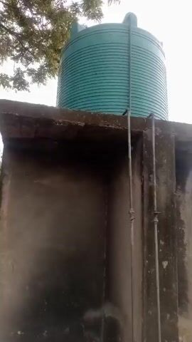 The video pans across an outdoor scene. Initially, a large green water tank is visible, elevated on a concrete structure. A metal ladder is attached to the side of the water tank. The camera then moves to show a yard with some scattered debris, trees, and buildings in the background. Further on, two trees stand in a sandy area, with a brick wall and buildings behind them. The scene shifts to a person in blue clothing tending to a garden bed near a building. The video concludes by showing the exterior of a building with barred windows and doors, and a paved walkway in front. The environment appears to be overcast.