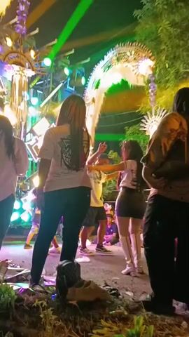 The video shows a group of people dancing at an outdoor event at night. Stage lights are projecting green and yellow beams onto the scene. The people are in the foreground and midground, with some appearing to be dancing energetically. In the background, there is what appears to be a stage setup with lights and decorative elements. The ground is uneven and appears to be dirt or gravel, with some debris visible.