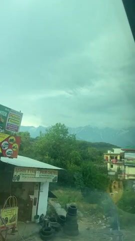 The video shows a view of mountains and foliage under a cloudy sky, seen through a window. The view includes a building with a red door and a billboard that displays a photograph of a temple. As the camera focuses upward, two birds fly into the shot from behind the building and head towards the mountains. The first bird flies out of frame and the second one remains, continuing its path. The scene is overcast, with the landscape appearing in shades of green. There's also a glimpse of a white van on the roadside.