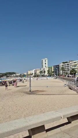 The video shows a sunny beach with many people. The sand is light brown, and there are numerous beach umbrellas of various colors, including blue, green, and yellow. People are spread out on the sand, some sitting under umbrellas, others walking or standing. In the background, there are several buildings, including a tall white building with many windows, and other multi-story structures. The sky is clear and blue. The water of the sea is visible on the left side of the frame, with a sailboat in the distance. A few people are walking on the beach, dressed in swimwear. There is also a metal shower fixture on the beach. The scene suggests a typical summer day at a crowded beach resort.
