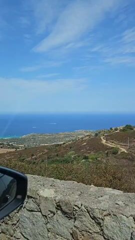 The video shows a view from a car looking out over a landscape with the ocean in the distance. In the foreground, a stone wall is visible. Beyond the wall is a hilly terrain with green vegetation and a dirt road winding through it. The ocean is a deep blue, and the sky is clear with a few scattered clouds. A car's side mirror is visible on the left side of the frame.