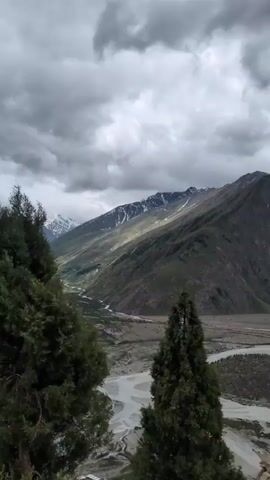 The video is a vertical, panning shot of a mountainous landscape under a cloudy sky. The camera moves from right to left, revealing a wide river flowing through a valley. The valley is flanked by steep, green mountains, some of which are capped with snow. The riverbed appears wide and braided with multiple channels. The vegetation on the mountainsides is sparse in some areas and more lush in others. There is a road visible along the side of the valley, with a yellow and black striped barrier in the foreground. The sky is filled with dramatic, dark clouds, suggesting a potential for rain or a storm. The overall impression is one of a vast, rugged, and natural landscape.