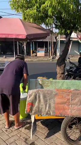 The video shows an outdoor scene with several people and objects. In the foreground, a part of a motorcycle is visible, with its wheel and exhaust. Behind it, there is a cart with a blue and yellow frame and a tarp-covered compartment. The cart is positioned on a sidewalk.

In the middle ground, a tree stands to the left, with its trunk visible. More of the sidewalk is visible, with some coconuts scattered around the base of the tree. Several people are present. One person, wearing a black t-shirt and a cap, is crouched down and appears to be working with coconuts. Another person, wearing a black t-shirt, is sitting on a motorcycle. Further back, another person is partially visible, wearing a light-colored headscarf.

The background reveals a street with a few buildings. The buildings have roofs and walls typical of residential or small commercial structures. The overall lighting suggests it is daytime, possibly late afternoon, with sunlight casting shadows. The scene appears to be in a public area, possibly a street market or a roadside stall.