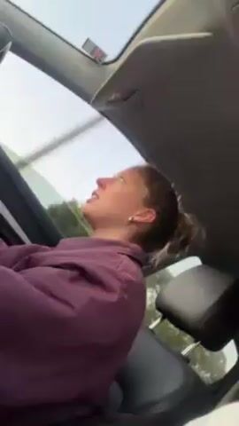 A woman is driving a car. She is wearing a maroon jacket and has her hair in a bun. The car has a sunroof. She is listening to music and makes a silly face at the camera, sticking out her tongue and widening her eyes.