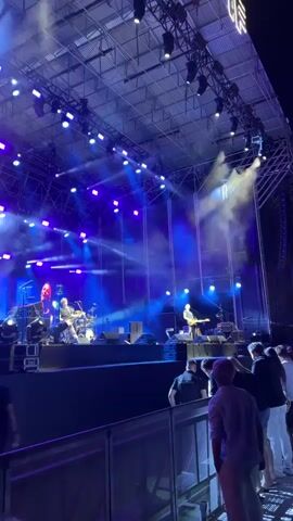 A concert is taking place on an outdoor stage. The stage is illuminated by blue and purple lights, with smoke effects in the air. Two musicians are visible on stage. One musician is seated at a drum kit, and another is standing and playing a guitar. Several audience members are visible in the foreground, facing the stage. The audience members appear to be standing behind a railing. A large white structure, possibly a tent or stage covering, is visible above the stage. The sky above is dark, indicating it is nighttime.