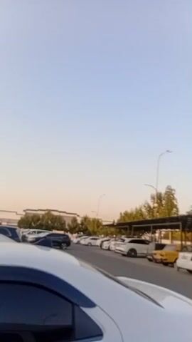 The video shows an outdoor parking lot with a covered area for cars. Several cars are parked under the canopy, including a white van, a black sedan, and a yellow car. The ground is asphalt, and there are trees and buildings in the background. The sky is clear and blue. The camera is moving, panning across the parking lot.