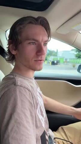 A young man with brown hair is sitting in the driver's seat of a car. He is wearing a tan t-shirt. The camera is positioned inside the car, focusing on the man's profile. He turns to look directly at the camera and smiles. The background outside the car window shows a storage facility and some trees. The video captures a candid moment where the man appears to be surprised by the filming.